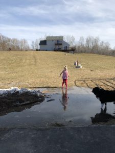 Little girl in pink jumping into large puddle at end of hill, with house on top of hill.